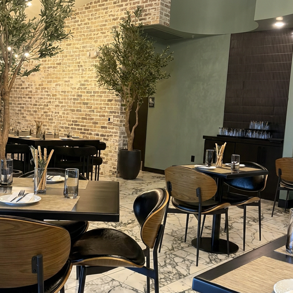Elegant restaurant dining room with set tables, striped banquette seating, olive trees, exposed brick, and marble flooring.