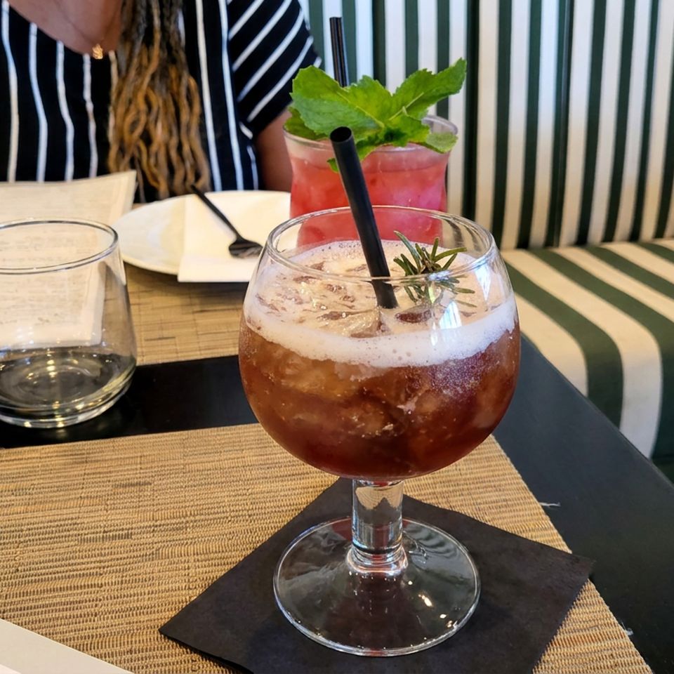 Cocktail with ice, foam, rosemary, and straw on a restaurant table, with a red mint-garnished drink and striped seating behind.